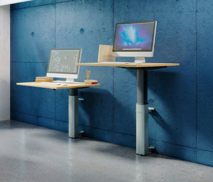 Two ConSet 501-19 wall-mounted desks shown at different heights against a textured blue concrete-style wall. One desk is at a sitting height and the other is at a standing height, each supporting a silver desktop computer. The desks feature light wood tops and silver-grey adjustable lifting columns, demonstrating the range of motion and space-saving design in a modern, industrial office setting.