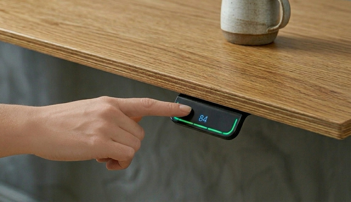 A close-up of a LINAK DPG1C desk controller mounted under a light oak wooden desktop. A hand is shown pressing the sleek, black digital interface which displays the number "84" in blue light and a glowing green status bar. A ceramic mug is partially visible on the desk surface in the background.