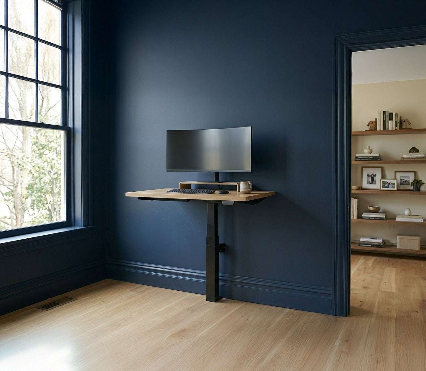 A minimalist single-leg LINAK wall-mounted standing desk with an oak wood top, installed against a dark navy blue wall. The desk features a curved ultra-wide monitor on a wooden riser, a black keyboard, and a mouse. The background shows a light wood floor and a built-in bookshelf, illustrating a space-saving home office design.