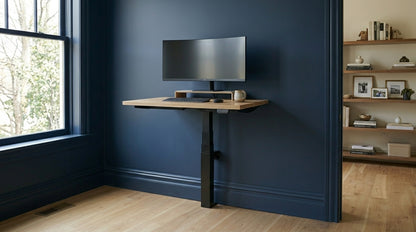 A minimalist single-leg LINAK wall-mounted standing desk with an oak wood top, installed against a dark navy blue wall. The desk features a curved ultra-wide monitor on a wooden riser, a black keyboard, and a mouse. The background shows a light wood floor and a built-in bookshelf, illustrating a space-saving home office design.