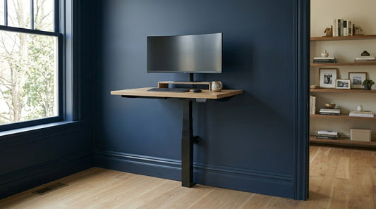 A minimalist single-leg LINAK wall-mounted standing desk with an oak wood top, installed against a dark navy blue wall. The desk features a curved ultra-wide monitor on a wooden riser, a black keyboard, and a mouse. The background shows a light wood floor and a built-in bookshelf, illustrating a space-saving home office design.