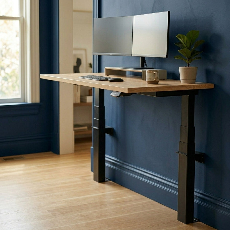 A wall-mounted sit-stand desk with a light oak desktop and black lifting columns, positioned against a dark navy blue accent wall. The workstation is styled with dual monitors on a riser, a wireless keyboard, a ceramic mug, and a small potted plant. The bright home office features light hardwood floors and a large window.